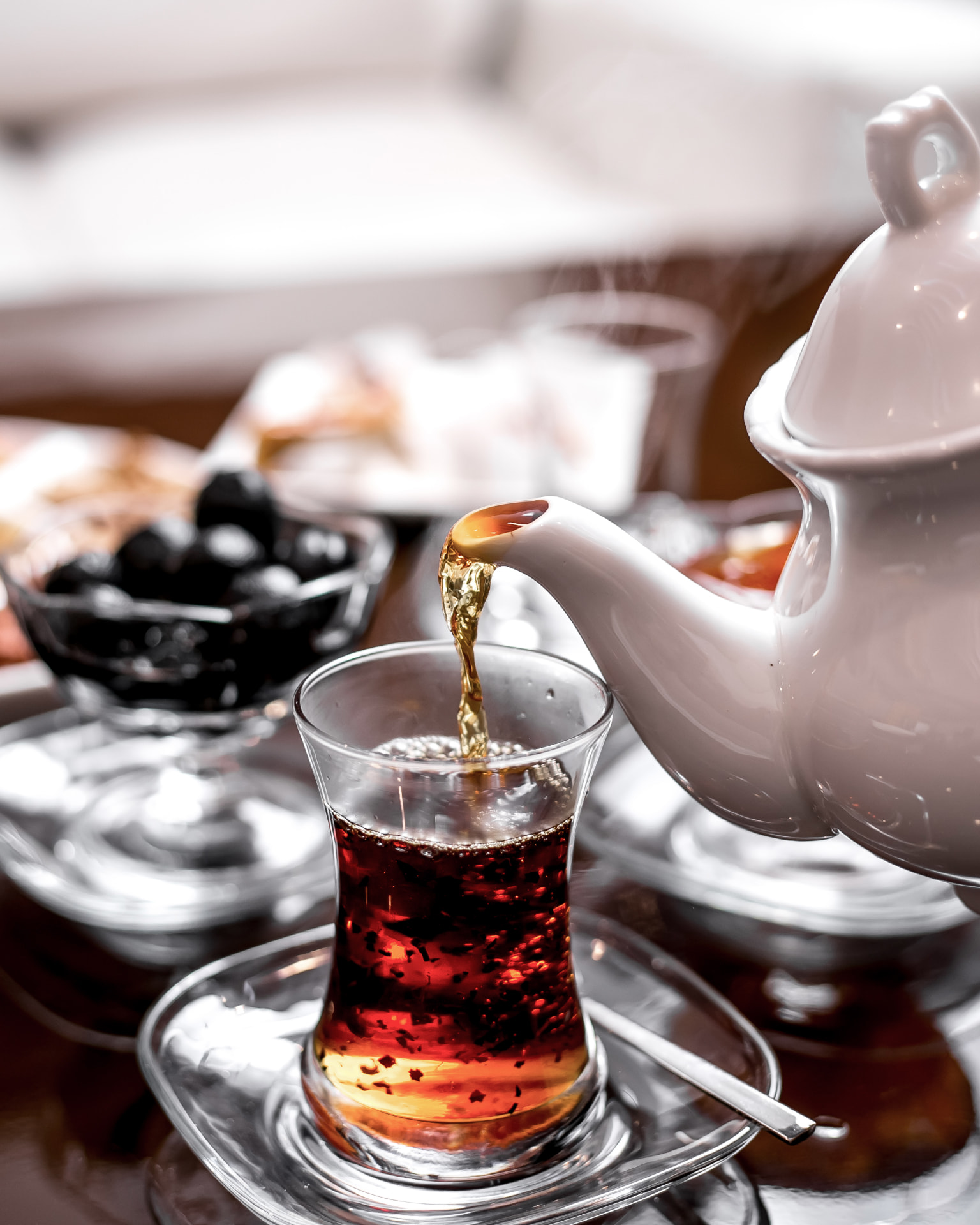 Tea poured from a white teapot into a glass armudu, with a warm table spread in the background